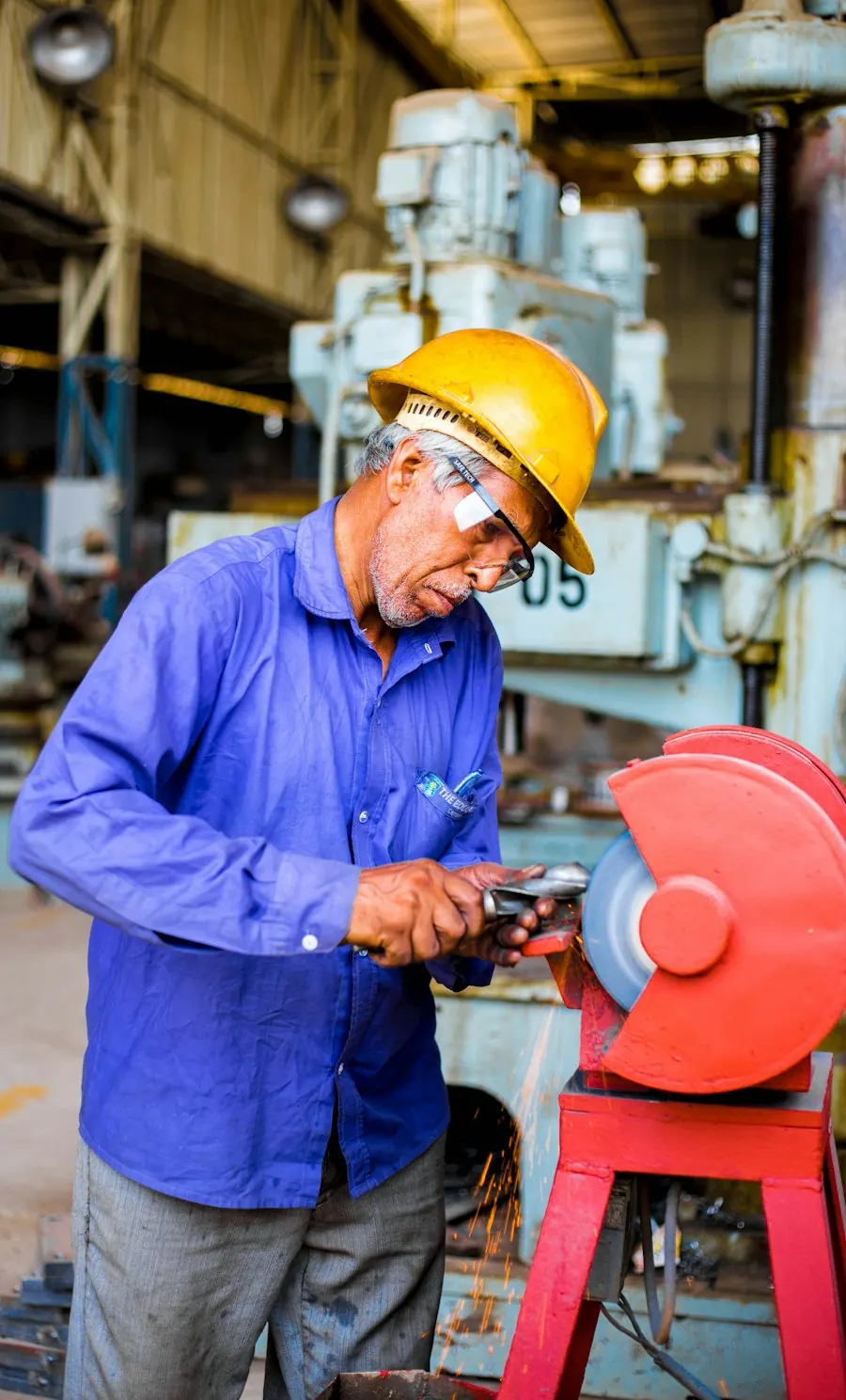 A worker in a yellow hard hat uses a bench grinder in a machine shop