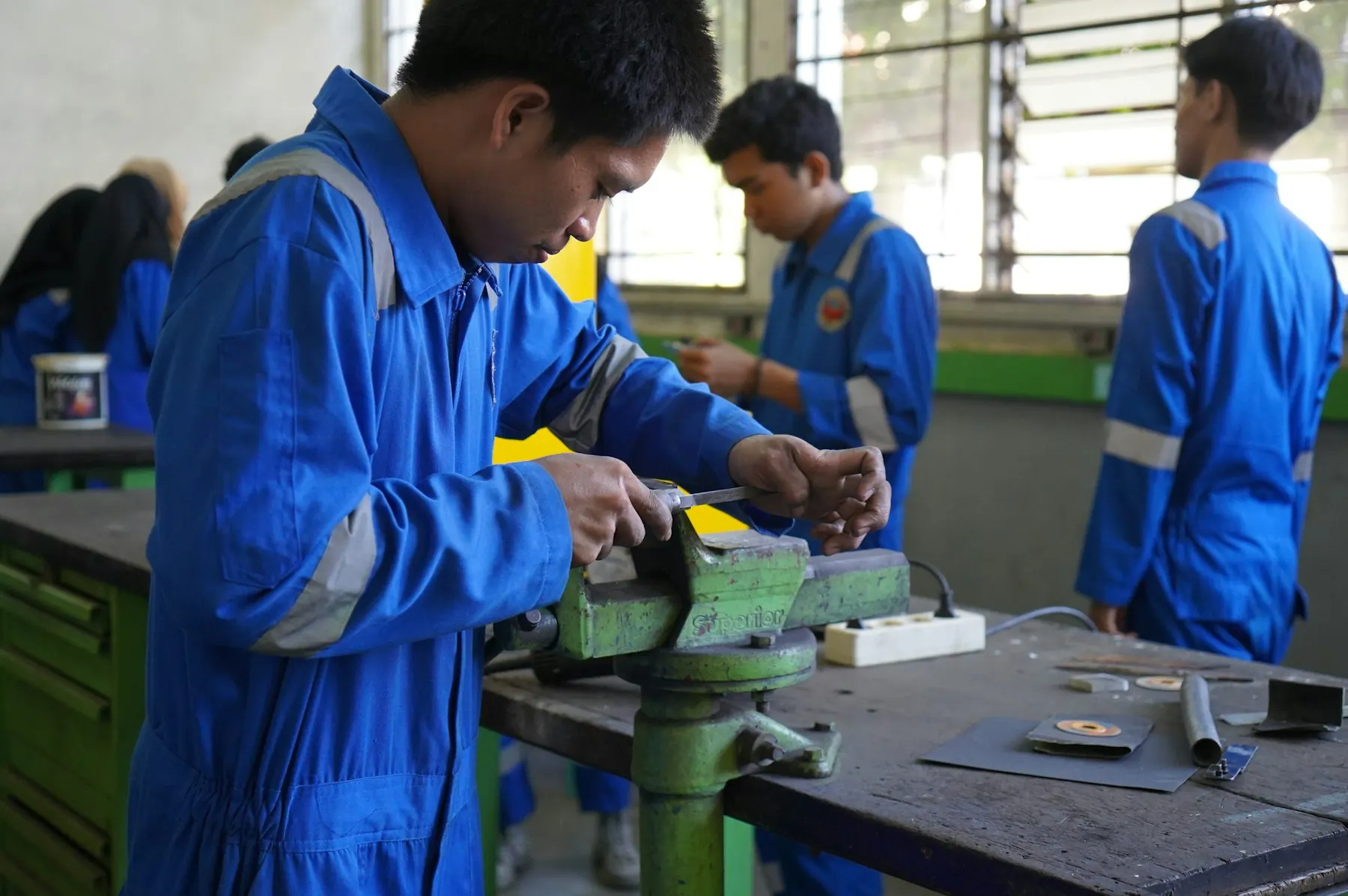 Trainees in blue coveralls work at a bench vise in a workshop