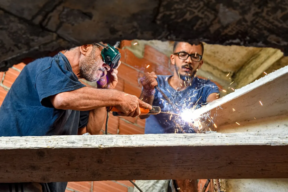 Two welders in protective gear work side by side on a joint, sparks flying